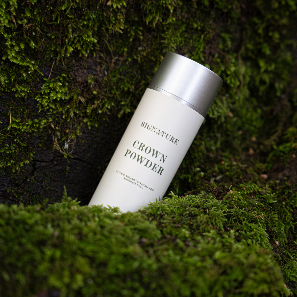A white cylindrical bottle of Signature Crown Powder with a metallic silver cap, placed diagonally on a bed of green moss against a dark, mossy background.