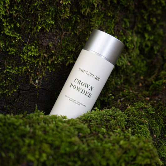 A white cylindrical bottle of Signature Crown Powder with a metallic silver cap, placed diagonally on a bed of green moss against a dark, mossy background.