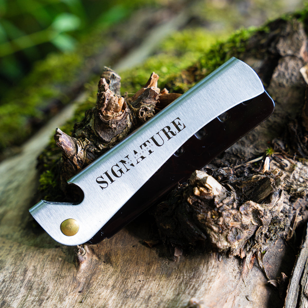 A folding pocket comb with a brushed silver metal blade and a black handle, resting on a piece of weathered wood outdoors. The comb is engraved with the word 'SIGNATURE'.