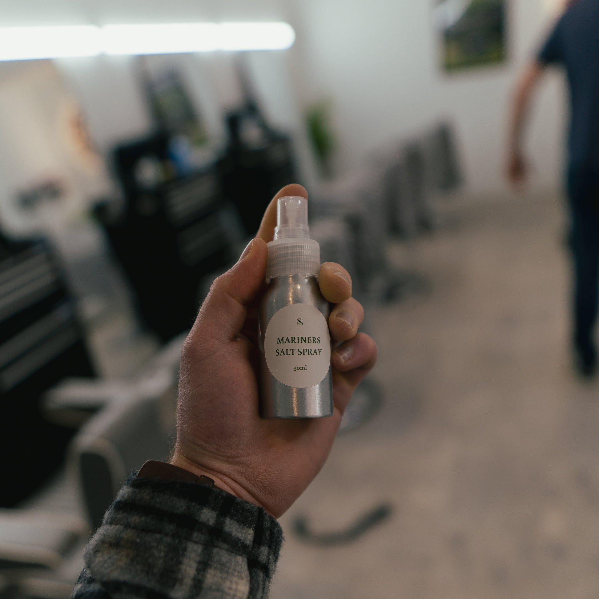 Hand holding a small spray bottle labeled 'Mariners Salt Spray' in a blurred salon setting.