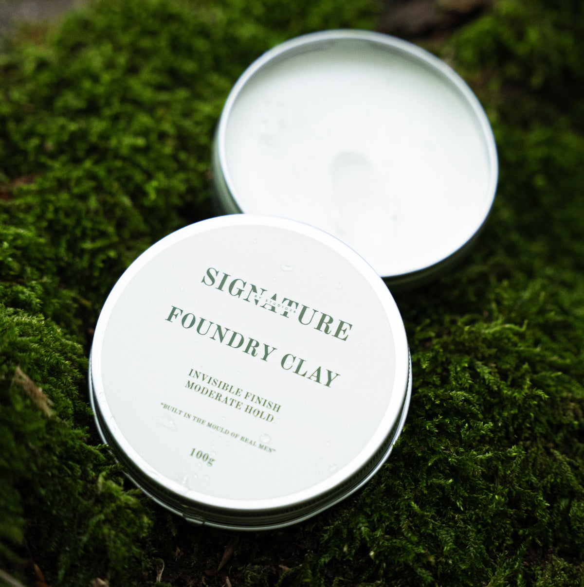 A round metal tin of Signature Foundry Clay hair product, shown open with the white cream visible inside, resting on a bed of green moss. The lid is placed in front, displaying the green printed brand name and product name.