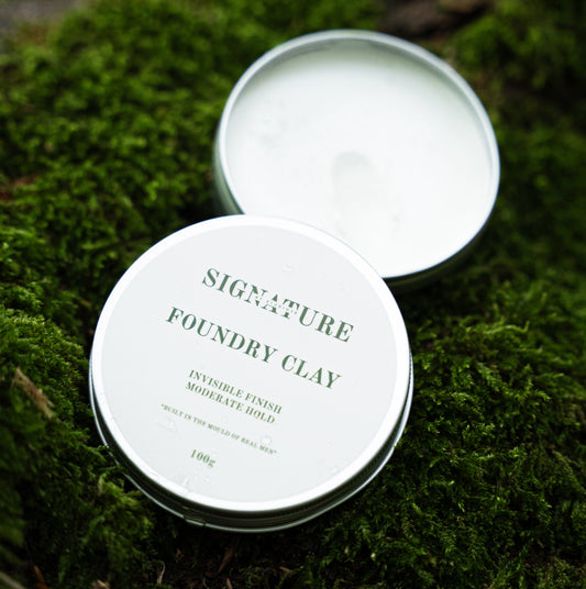 A round metal tin of Signature Foundry Clay hair product, shown open with the white cream visible inside, resting on a bed of green moss. The lid is placed in front, displaying the green printed brand name and product name.