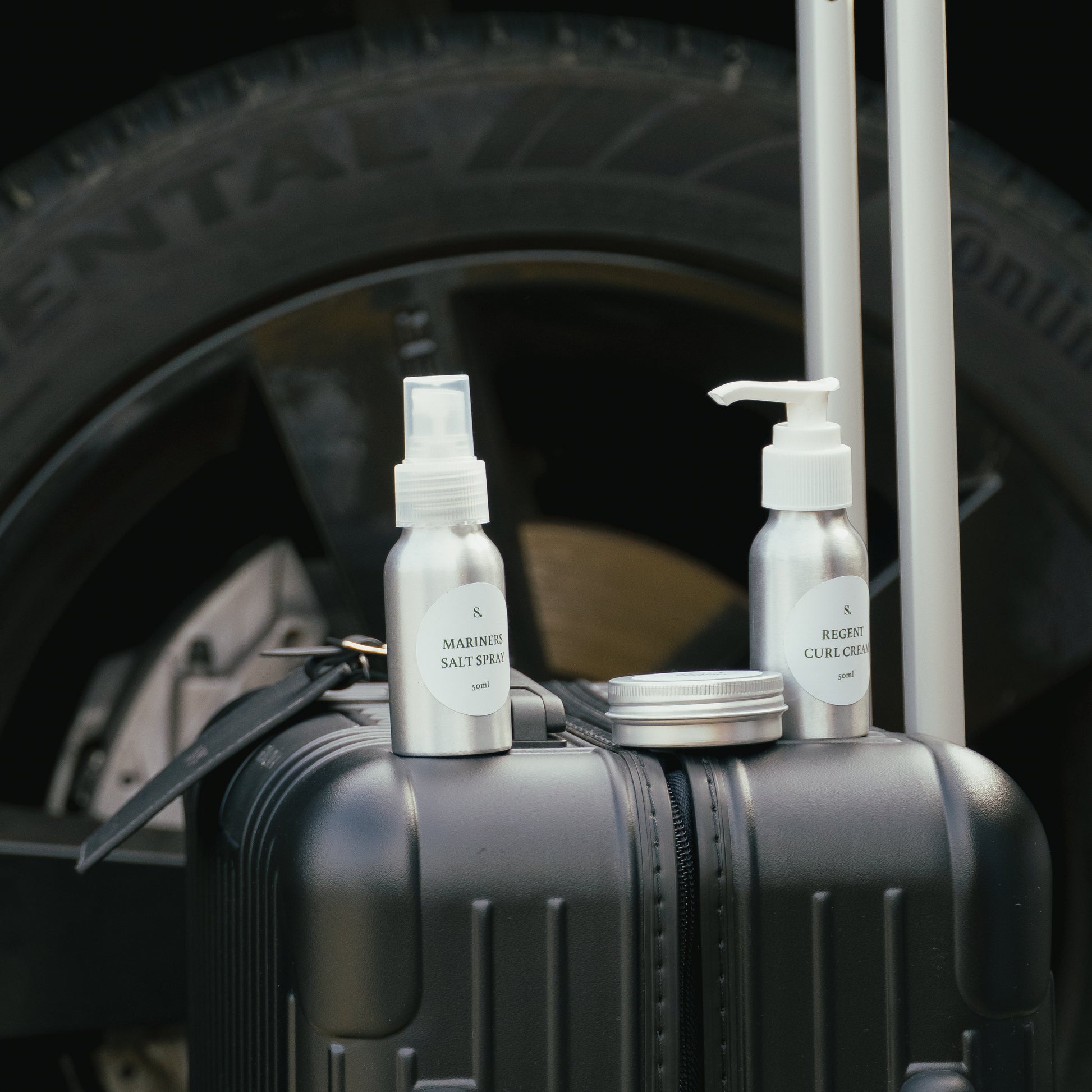 Three hair styling travel bottles on a suitcase with a car tire in the background