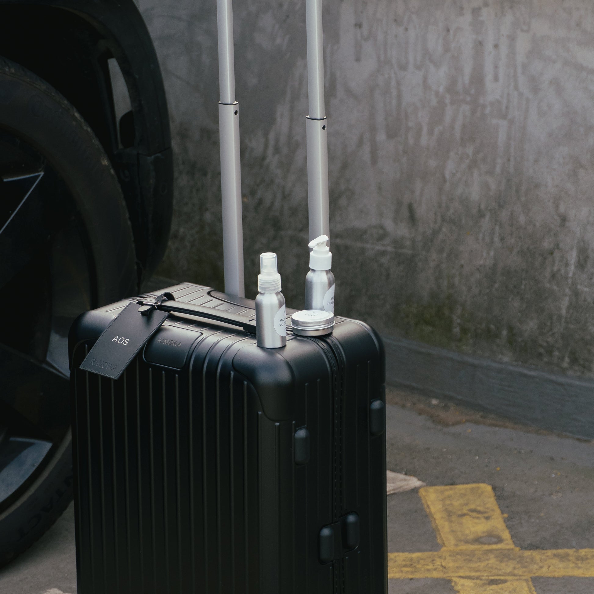 Black suitcase with a tag and three travel bottles on a concrete surface next to a vehicle.