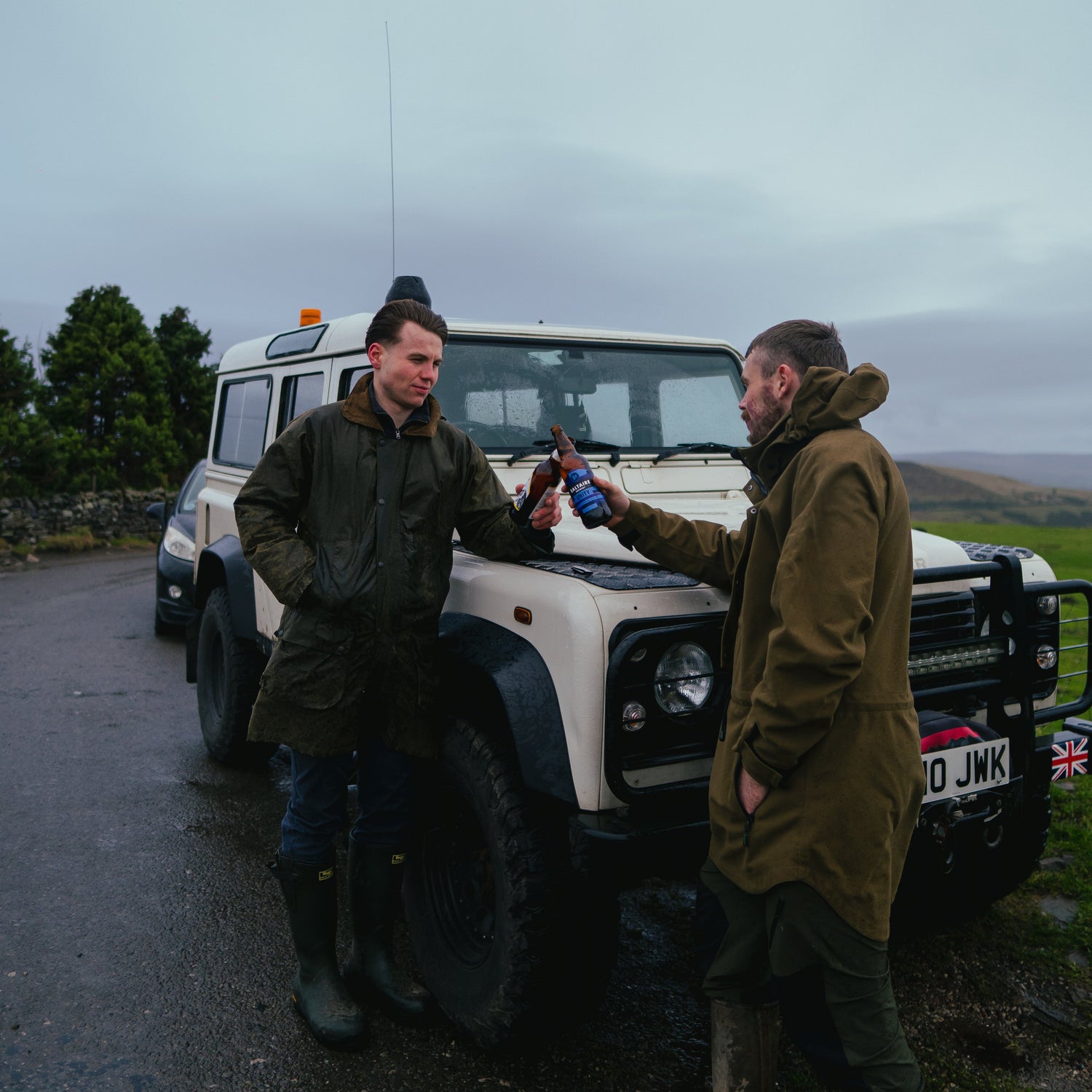 Two men standing next to a white Land Rover on a road with a scenic background.