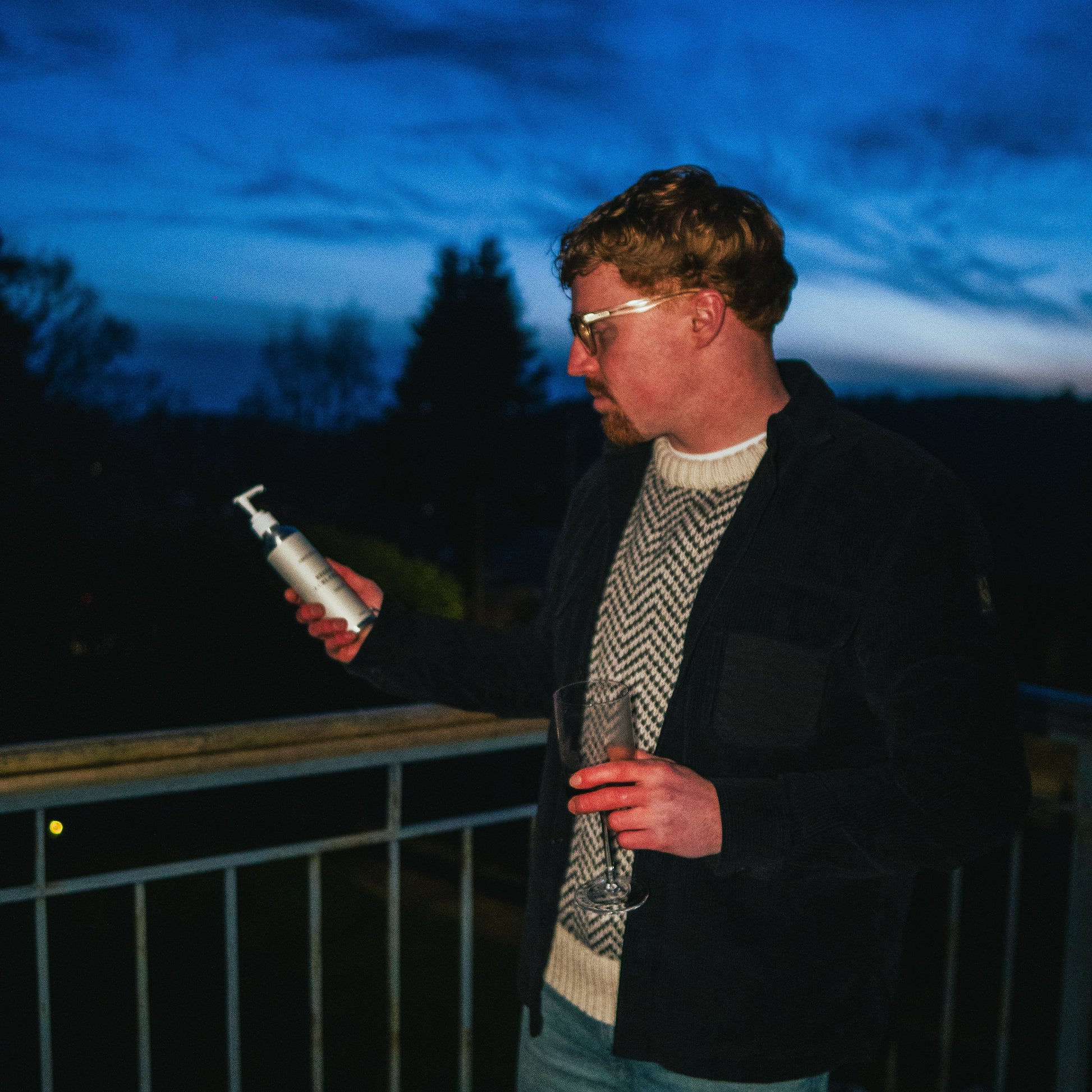 Man on a balcony at dusk holding a curl cream, with a dark sky and trees in the background.