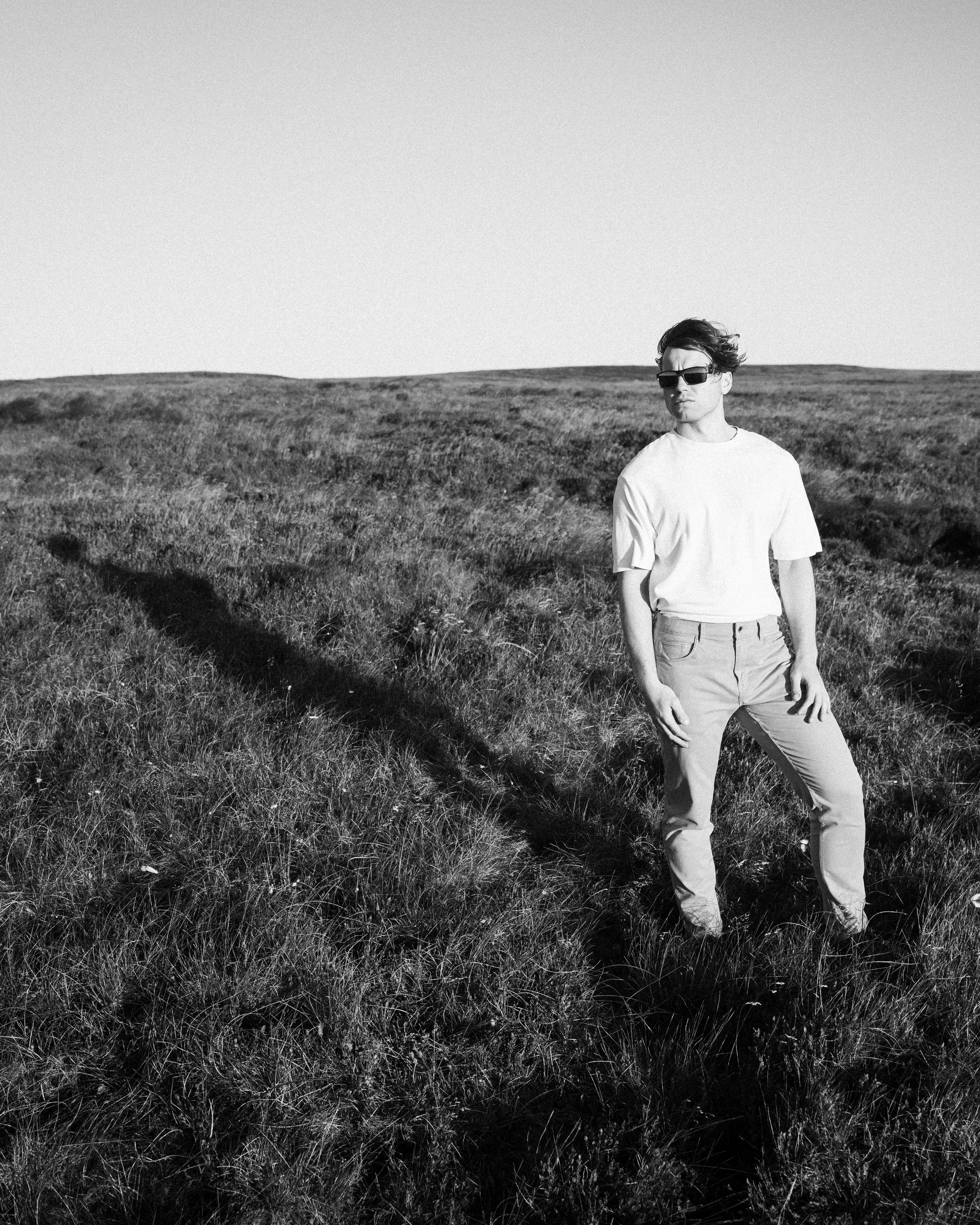 A black and white photograph of a man wearing sunglasses, a white t-shirt, and light-colored trousers standing in a grassy field under a clear sky.