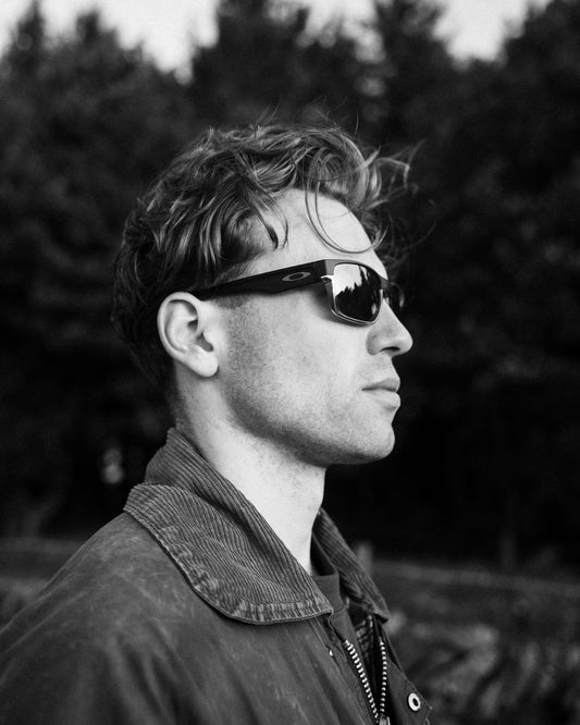 A black and white portrait of a man in profile wearing dark sunglasses and a corduroy jacket, with a blurred outdoor background of trees.