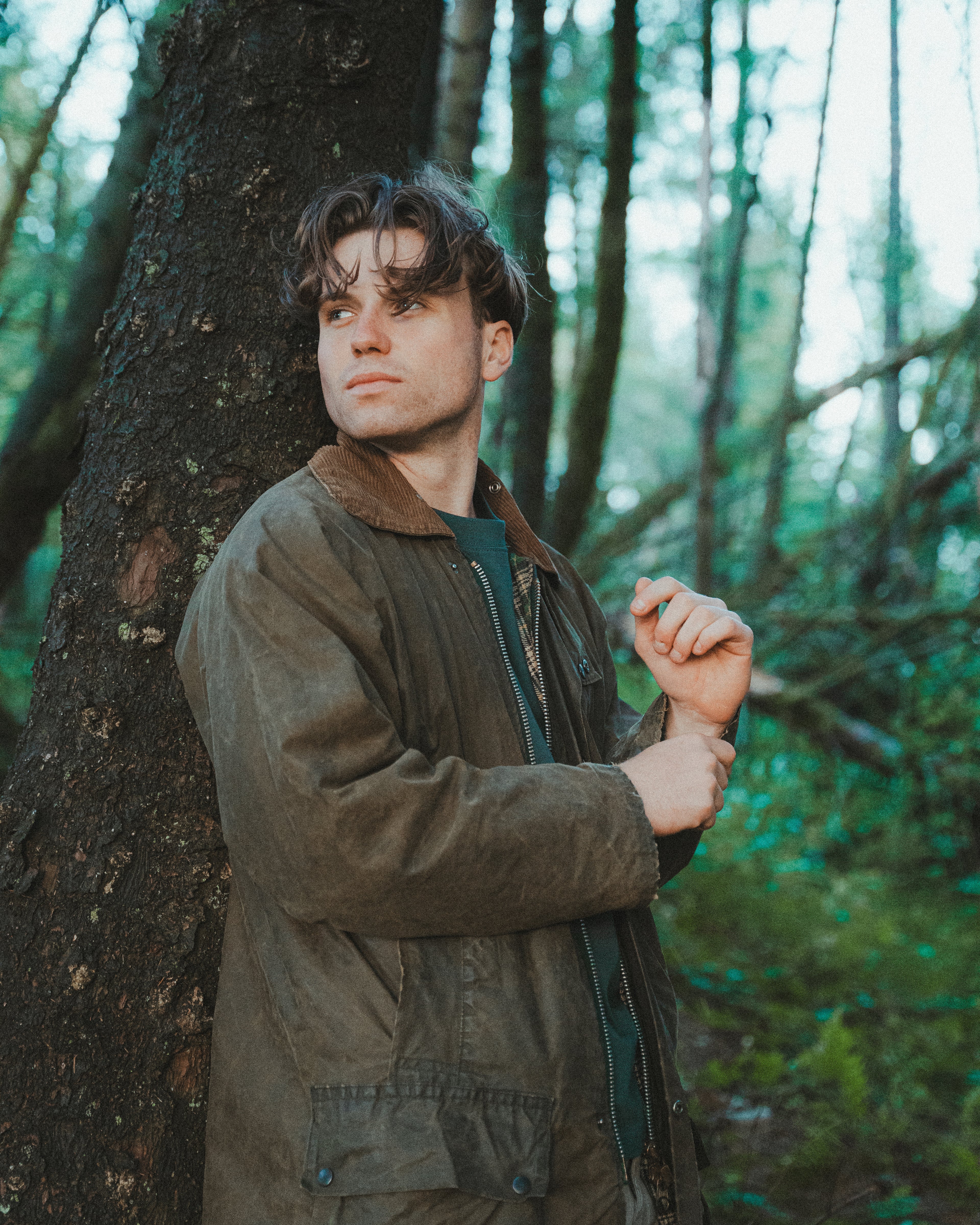 A man wearing a brown waxed canvas utility jacket with a brown corduroy collar, standing in a forest next to a large tree trunk.