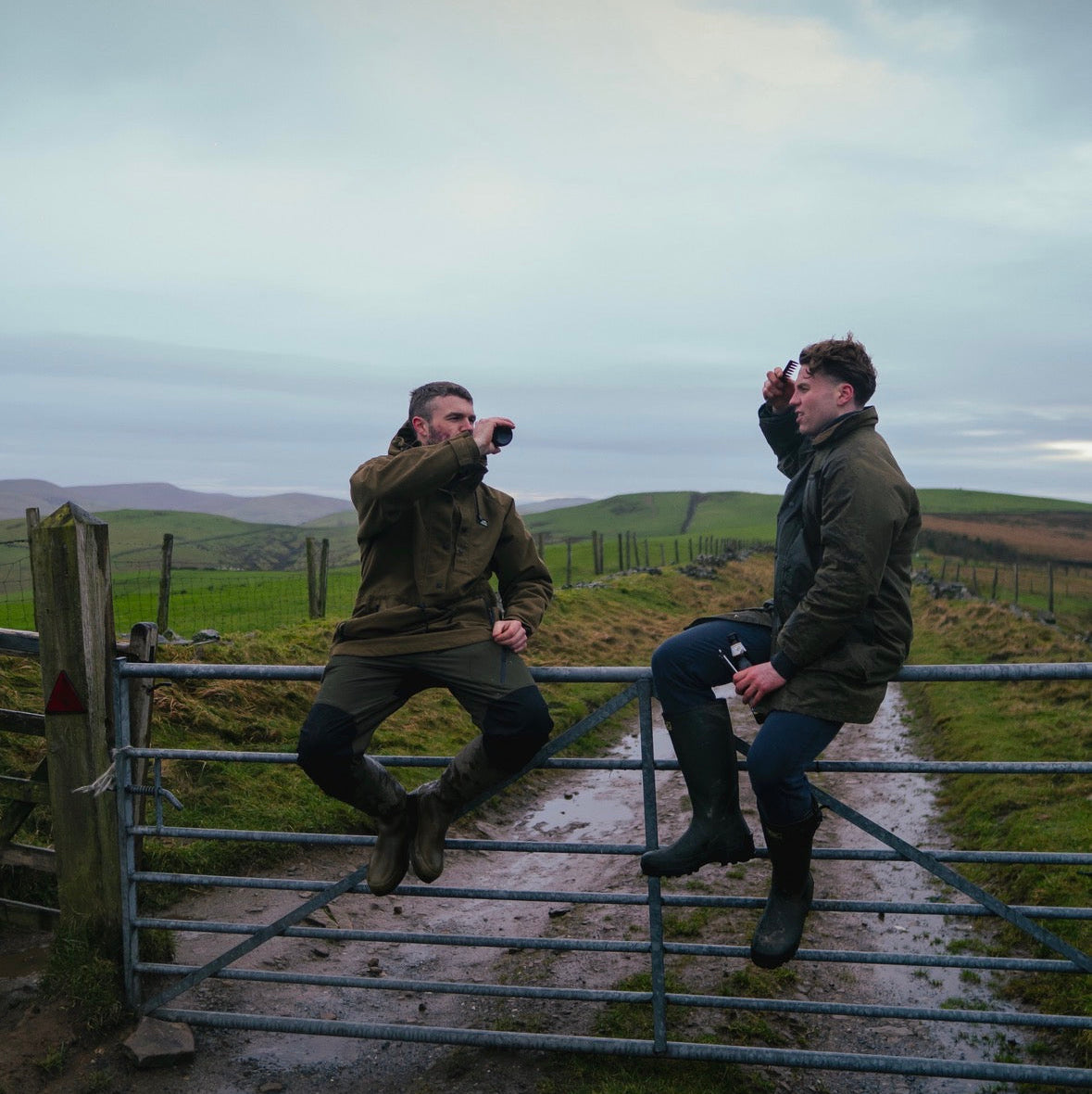Two people sitting on a gate in a rural landscape with a cloudy sky.